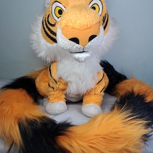 May include: A plush tiger toy with orange and black striped fur, a white chest, and white muzzle. The tiger has large yellow eyes with black pupils, black ear tips, and a long, thick, fluffy tail with alternating orange and black fur sections.