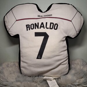 Puede incluir: Un coj&iacute;n blanco con forma de camiseta que presenta bordes negros y el texto REAL MADRID. El nombre RONALDO y el n&uacute;mero 7 est&aacute;n impresos en negro en la parte frontal. Un peque&ntilde;o logotipo circular es visible en la base del n&uacute;mero 7 del coj&iacute;n.