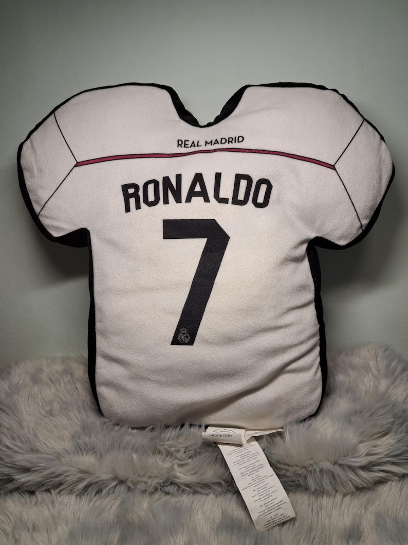 Puede incluir: Un coj&iacute;n blanco con forma de camiseta que presenta bordes negros y el texto REAL MADRID. El nombre RONALDO y el n&uacute;mero 7 est&aacute;n impresos en negro en la parte frontal. Un peque&ntilde;o logotipo circular es visible en la base del n&uacute;mero 7 del coj&iacute;n.