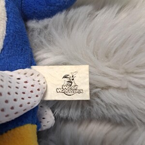 May include: A plush toy featuring blue, yellow, and white fabric sections. A white fabric tag is attached, displaying a black graphic illustration of the character Woody Woodpecker along with the text Woody Woodpecker printed in black ink.