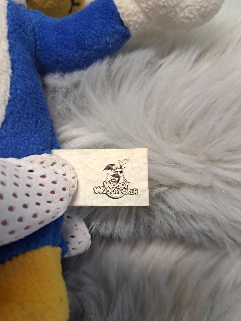 May include: A plush toy featuring blue, yellow, and white fabric sections. A white fabric tag is attached, displaying a black graphic illustration of the character Woody Woodpecker along with the text Woody Woodpecker printed in black ink.