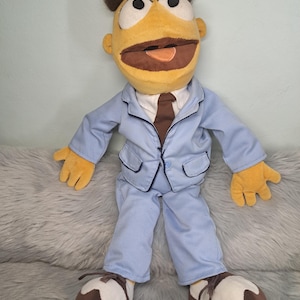 May include: A plush puppet doll with yellow skin, brown hair, and large white eyes. The doll wears a light blue suit jacket with dark blue piping, matching light blue trousers, a white collared shirt, a brown necktie, and brown and white shoes.