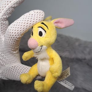 May include: A small yellow plush toy of the character Roo from Winnie the Pooh, featuring a pink nose, pink-lined ears, a white chest patch, and embroidered facial features. A white fabric tag is attached to the side with printed text.