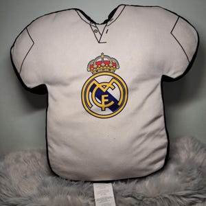 Puede incluir: Un coj&iacute;n de peluche blanco con forma de camiseta que presenta el logotipo del club de f&uacute;tbol Real Madrid en la parte delantera. El logo incluye una corona dorada, un emblema circular azul y dorado con las letras MCF y un borde negro.