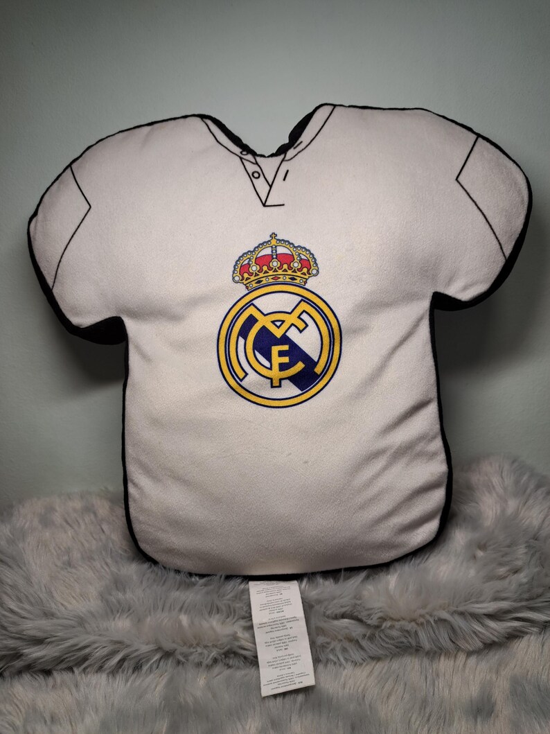 Puede incluir: Un coj&iacute;n de peluche blanco con forma de camiseta que presenta el logotipo del club de f&uacute;tbol Real Madrid en la parte delantera. El logo incluye una corona dorada, un emblema circular azul y dorado con las letras MCF y un borde negro.
