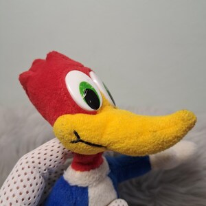 May include: A plush toy of the cartoon character Woody Woodpecker, featuring a bright red crest, a long yellow beak, large white eyes with black pupils, and a blue body with a white chest patch, captured in a side profile view.