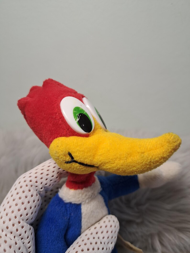May include: A plush toy of the cartoon character Woody Woodpecker, featuring a bright red crest, a long yellow beak, large white eyes with black pupils, and a blue body with a white chest patch, captured in a side profile view.