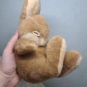 May include: A plush, brown bunny rabbit toy with tan accents on the ears, paws, and belly. The stuffed animal is soft and fluffy, with a small, closed eye. The bunny is held in a person's hand.