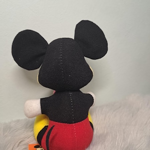 Pu&ograve; includere: Un pupazzo di peluche di Topolino, visto da dietro, con orecchie nere, un top nero, pantaloncini rossi e una base gialla e nera. La bambola ha guanti bianchi e un'etichetta con una grafica di Topolino.