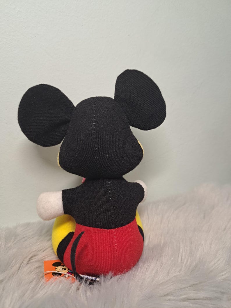 Pu&ograve; includere: Un pupazzo di peluche di Topolino, visto da dietro, con orecchie nere, un top nero, pantaloncini rossi e una base gialla e nera. La bambola ha guanti bianchi e un'etichetta con una grafica di Topolino.