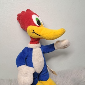 May include: A plush toy of the cartoon character Woody Woodpecker, featuring a bright red crest, a long yellow beak, large white eyes with green pupils, a blue body with a white chest, white hands, and yellow feet, standing in a profile pose.