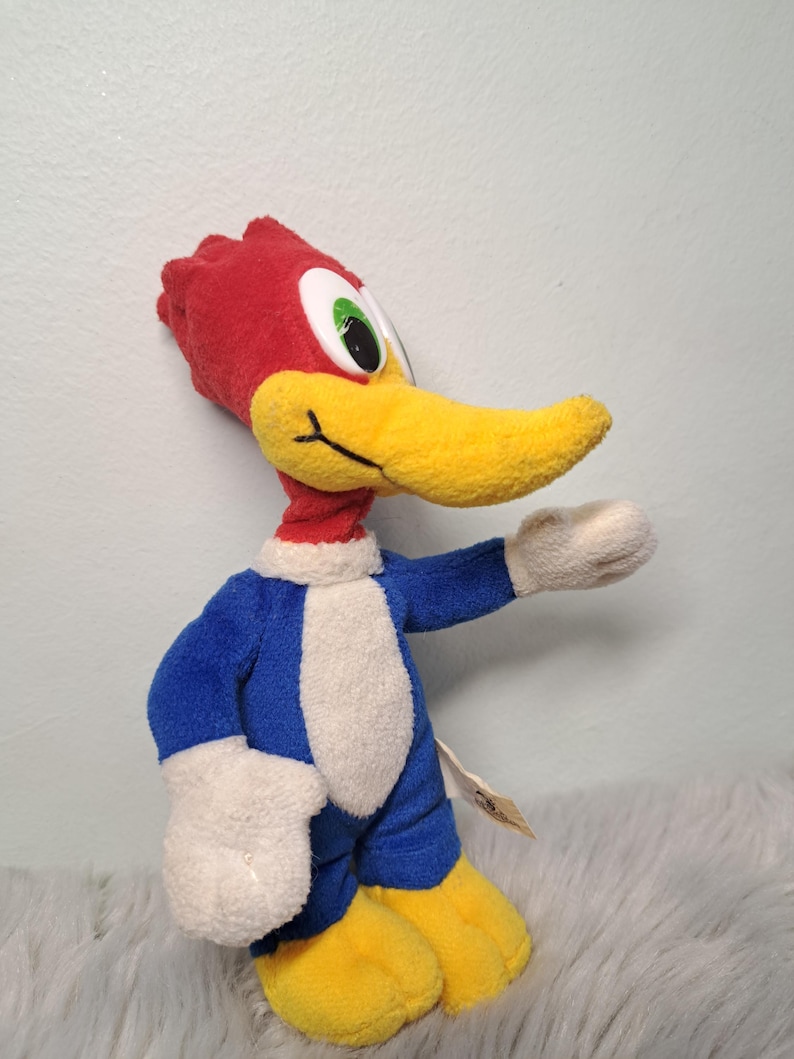 May include: A plush toy of the cartoon character Woody Woodpecker, featuring a bright red crest, a long yellow beak, large white eyes with green pupils, a blue body with a white chest, white hands, and yellow feet, standing in a profile pose.