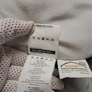 Puede incluir: Una etiqueta de cuidado de tela blanca unida a un art&iacute;culo textil blanco y suave. La etiqueta presenta el texto MADE IN CHINA, el logotipo de SkyBrands y s&iacute;mbolos de cuidado. Una etiqueta secundaria muestra el logotipo Oeko-Tex Standard 100.