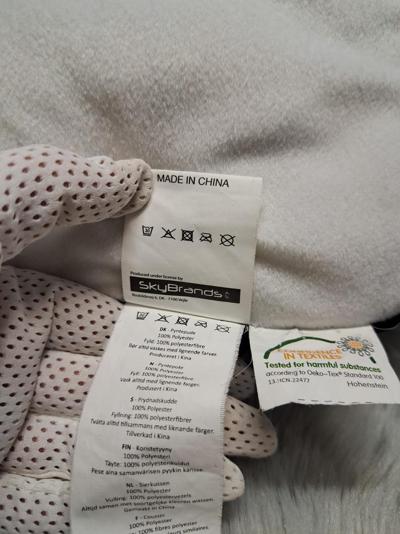 Puede incluir: Una etiqueta de cuidado de tela blanca unida a un art&iacute;culo textil blanco y suave. La etiqueta presenta el texto MADE IN CHINA, el logotipo de SkyBrands y s&iacute;mbolos de cuidado. Una etiqueta secundaria muestra el logotipo Oeko-Tex Standard 100.