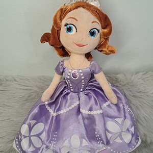 May include: A plush doll of a character with brown hair, blue eyes, and a silver tiara. The doll wears a purple satin dress featuring a white floral pattern along the hem, a printed bodice design, and a small purple amulet necklace around the neck.