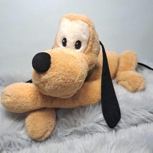 May include: A plush toy of Pluto, a cartoon dog character. The toy is light brown with a white face, black ears, and a black nose. It is lying down on a gray, fluffy surface.