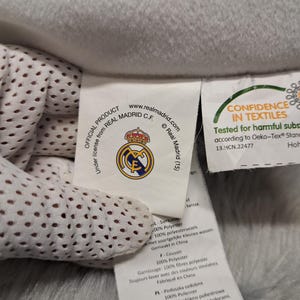 Puede incluir: Una etiqueta de tela blanca unida a un art&iacute;culo de forro polar gris claro. La etiqueta muestra el logotipo del Real Madrid C.F., el texto Official Product under license from Real Madrid C.F. y una etiqueta de certificaci&oacute;n Oeko-Tex.