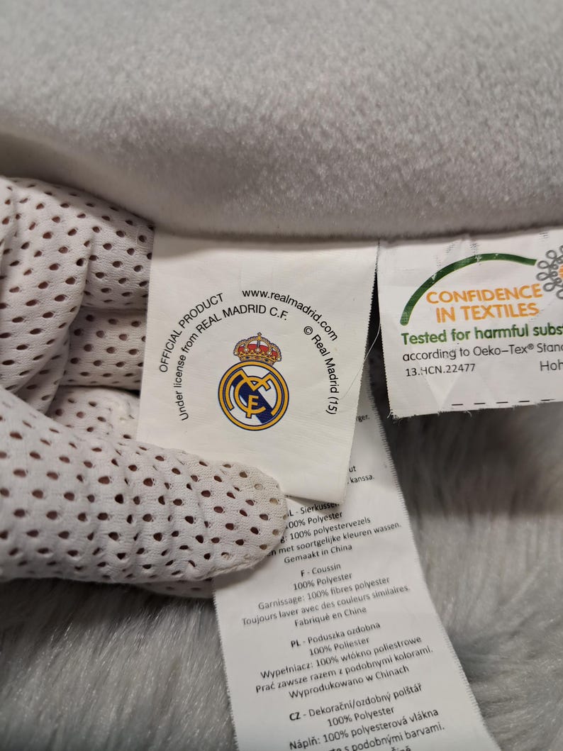 Puede incluir: Una etiqueta de tela blanca unida a un art&iacute;culo de forro polar gris claro. La etiqueta muestra el logotipo del Real Madrid C.F., el texto Official Product under license from Real Madrid C.F. y una etiqueta de certificaci&oacute;n Oeko-Tex.