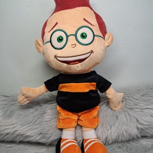 May include: A plush doll featuring a character with short red hair, green-rimmed glasses, and a wide smile. The doll wears a black t-shirt with an orange horizontal stripe, orange shorts, white socks, and orange sneakers with white stripes.