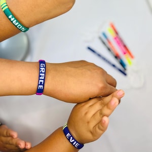 May include: Three personalised bracelets are displayed, each embroidered with a name. One is green with "NOAH", another is blue with "GRACE", and the third is blue with "EVIE". The bracelets are likely handmade.