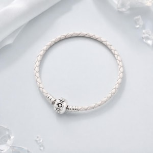 May include: A white braided leather bracelet featuring a cylindrical silver-toned metal charm. The charm is engraved with the text DORA and includes a small crown symbol above the letters. The bracelet has a smooth, circular, woven texture.