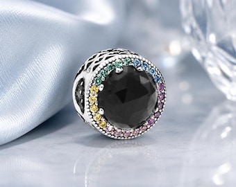 May include: A round silver charm bead featuring a large, faceted black center stone surrounded by a halo of small, multicolored gemstones in shades of yellow, green, blue, and pink. The metal frame has an intricate, openwork lattice pattern.