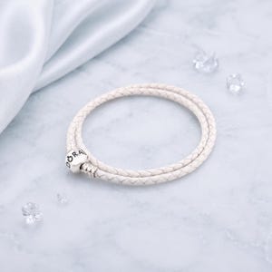 Puede incluir: Una pulsera de doble vuelta con un cordón de cuero trenzado blanco y un cierre cilíndrico de metal plateado. El cierre tiene grabado el texto DORA en letras negras. La pulsera está dispuesta en forma circular sobre una superficie plana.