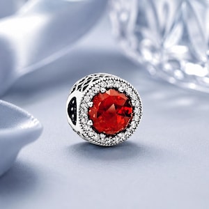 May include: A round charm bead featuring a large, faceted, circular red stone at the center, surrounded by a halo of small, clear, sparkling stones. The outer edge of the silver-toned metal bead displays an intricate, openwork lattice pattern.