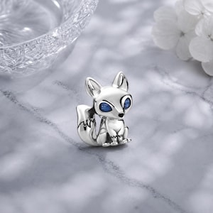 Peut inclure: Un charm en forme de renard argenté avec une finition métallique polie et deux petites pierres bleu foncé incrustées en guise d'yeux. Le renard est représenté assis avec la queue recourbée vers le haut, révélant des lignes gravées détaillées.