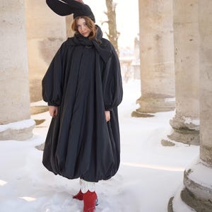 May include: A black, voluminous, long-sleeved coat with a gathered hem, worn with a matching black headpiece featuring two large, upright, rabbit-like ears with pink inner linings, a black scarf, and bright red boots with white ruffled cuffs.