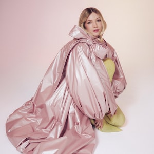 May include: A voluminous, metallic light pink coat featuring oversized sleeves and a large, prominent bow detail at the neckline. The garment is worn over a pair of textured, pale yellow leggings that extend down to the feet, creating a layered look.