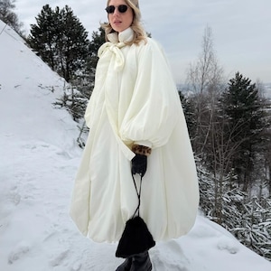 Peut inclure: Un manteau bouffant surdimensionné de couleur crème doté d'un col montant avec fermeture à nouer, de manches longues volumineuses et d'un ourlet arrondi. Il est associé à des gants en cuir noir, un petit sac à main en fourrure noire et des bottes.