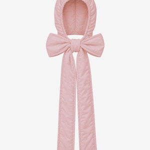 Puede incluir: Una capucha acolchada de color rosa claro con un lazo grande y estructurado en el cuello y dos cintas de tela rectangulares y largas que cuelgan hacia abajo. La textura acolchada es visible en toda la pieza, hecha de un material rosa pastel suave y uniforme.
