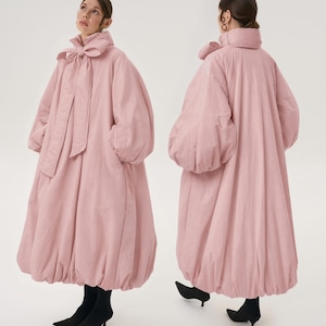 Puede incluir: Un vestido acolchado de manga larga y largo hasta los tobillos en un tono rosa pastel suave. La prenda presenta un cuello alto con un gran lazo de tela integrado en el cuello, mangas abullonadas voluminosas y un dobladillo fruncido y redondeado.