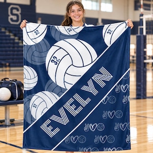 May include: A navy blue and white fleece blanket featuring a repeating pattern of volleyballs with the number 32, the name EVELYN in large white block letters, and small white line-art icons of volleyballs, hearts, and peace signs.