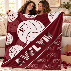 May include: A maroon and white fleece blanket featuring a repeating pattern of volleyballs with the number 32, the name EVELYN in large white block letters, and a smaller pattern of peace signs, hearts, and volleyballs along the bottom edge.