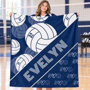 May include: A navy blue and white fleece blanket featuring a repeating pattern of volleyballs with the number 32, the name EVELYN in large white block letters, and a smaller pattern of peace signs, hearts, and volleyballs on the lower section.