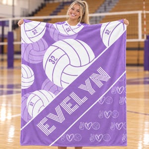 May include: A purple and white fleece blanket featuring a repeating pattern of volleyballs with the number 32 printed on them. A diagonal white stripe across the centre displays the name EVELYN in bold, white, striped, capital letters.