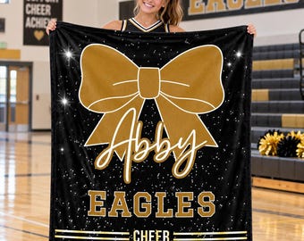 May include: A black fleece blanket featuring a large gold cheer bow graphic, the name Abby in white script, and the text EAGLES and CHEER in gold block letters. The design includes a white speckled pattern across the black background.