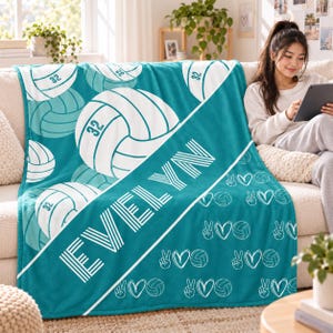 May include: A teal fleece blanket featuring a repeating pattern of white volleyballs with the number 32, a diagonal white stripe with the name EVELYN in white block letters, and a smaller pattern of peace signs, hearts, and volleyballs.