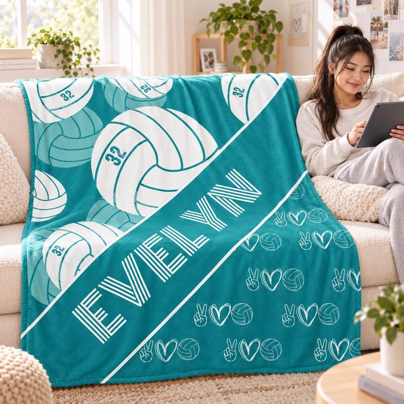 May include: A teal fleece blanket featuring a repeating pattern of white volleyballs with the number 32, a diagonal white stripe with the name EVELYN in white block letters, and a smaller pattern of peace signs, hearts, and volleyballs.