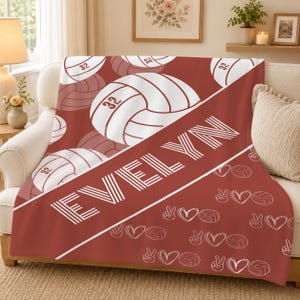 May include: A soft throw blanket in a deep terracotta red colour featuring a repeating pattern of white volleyballs with the number 32, the name EVELYN in bold white capital letters, and a scattered design of small white hearts and peace signs.