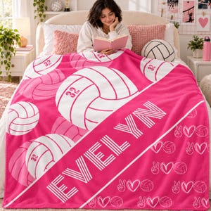 May include: A bright pink fleece blanket featuring a repeating pattern of white volleyballs, each marked with the number 32. A diagonal white stripe across the centre displays the name EVELYN in bold, white, block-style lettering.