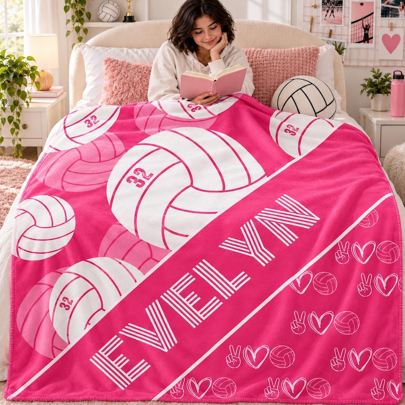 May include: A bright pink fleece blanket featuring a repeating pattern of white volleyballs, each marked with the number 32. A diagonal white stripe across the centre displays the name EVELYN in bold, white, block-style lettering.