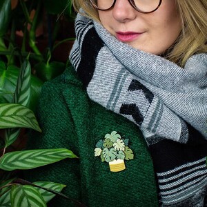 May include: A brooch depicting a potted plant, with a white pot and yellow woven detail, and green leaves. The brooch is pinned to a dark green coat. The person is wearing a grey and black scarf.