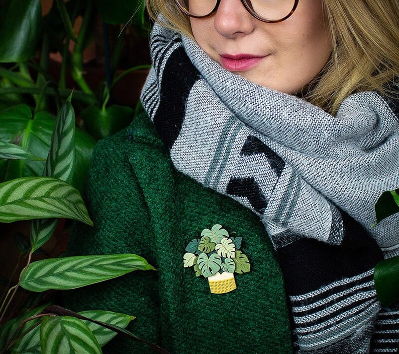 May include: A brooch depicting a potted plant, with a white pot and yellow woven detail, and green leaves. The brooch is pinned to a dark green coat. The person is wearing a grey and black scarf.
