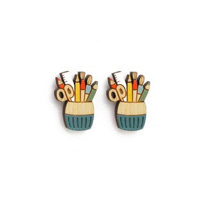 May include: A pair of wooden earrings shaped like a pencil holder filled with colorful pencils, crayons, and a ruler. The pencil holder is painted a light blue color with a white rim.