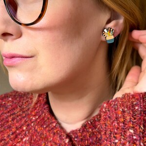 May include: A pair of earrings featuring a small, blue, and yellow enamel design of a pencil holder with pencils and paintbrushes sticking out of it.