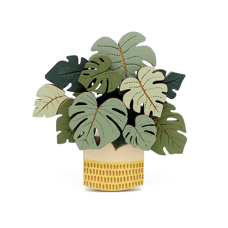 May include: A wooden decorative plant with large, green monstera leaves in a cream-coloured pot with a yellow dotted pattern. The leaves vary in shades of green and have brown vein details.
