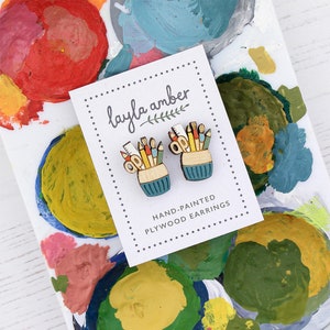 May include: A pair of hand-painted wooden earrings shaped like a pencil holder filled with colorful art supplies. The earrings are teal blue with a yellow and brown pencil holder. The text on the card reads "layla amber" and "HAND-PAINTED PLYWOOD EARRINGS".